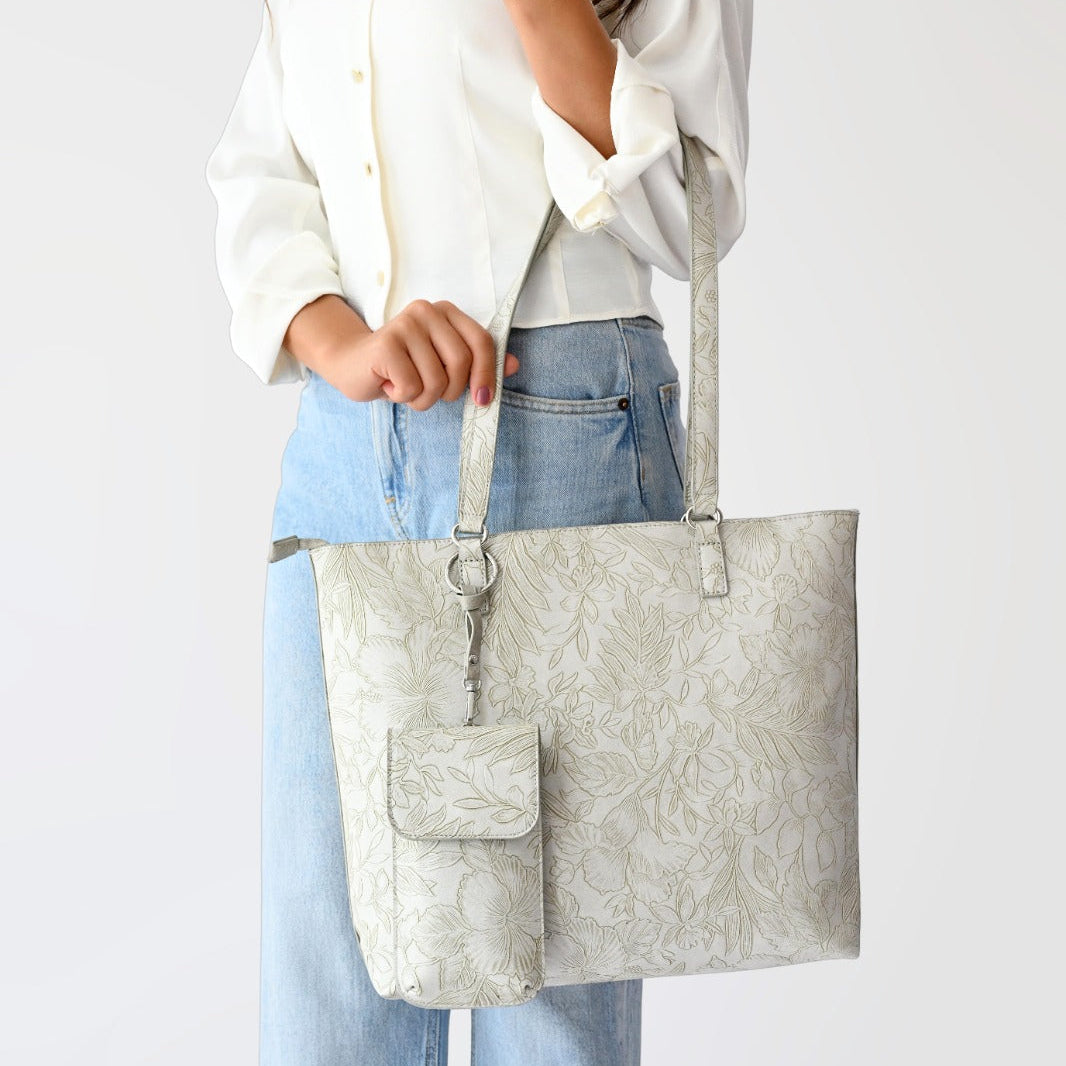 Woman wearing a white button-up shirt and light blue jeans carrying a cream floral jacquard tote bag with matching pouch and keychain.